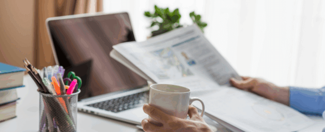 Professional reading morning business news with coffee at a home office desk with laptop and stationery.