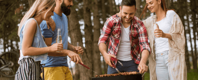 Four friends gathered around a BBQ grill, smiling and cooking skewers while holding drinks, symbolising casual BBQ tax myth business conversations.