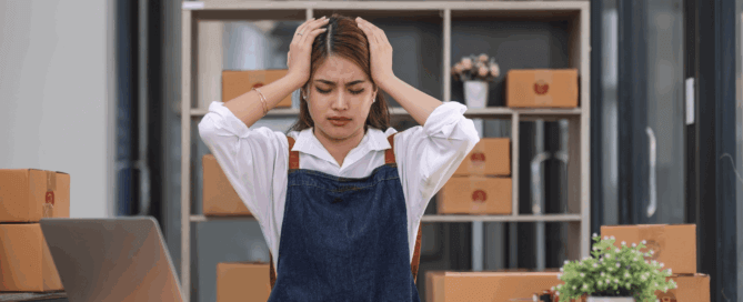 Small business owner looking stressed while working on finances at a desk with a laptop, calculator, and boxes in the background.